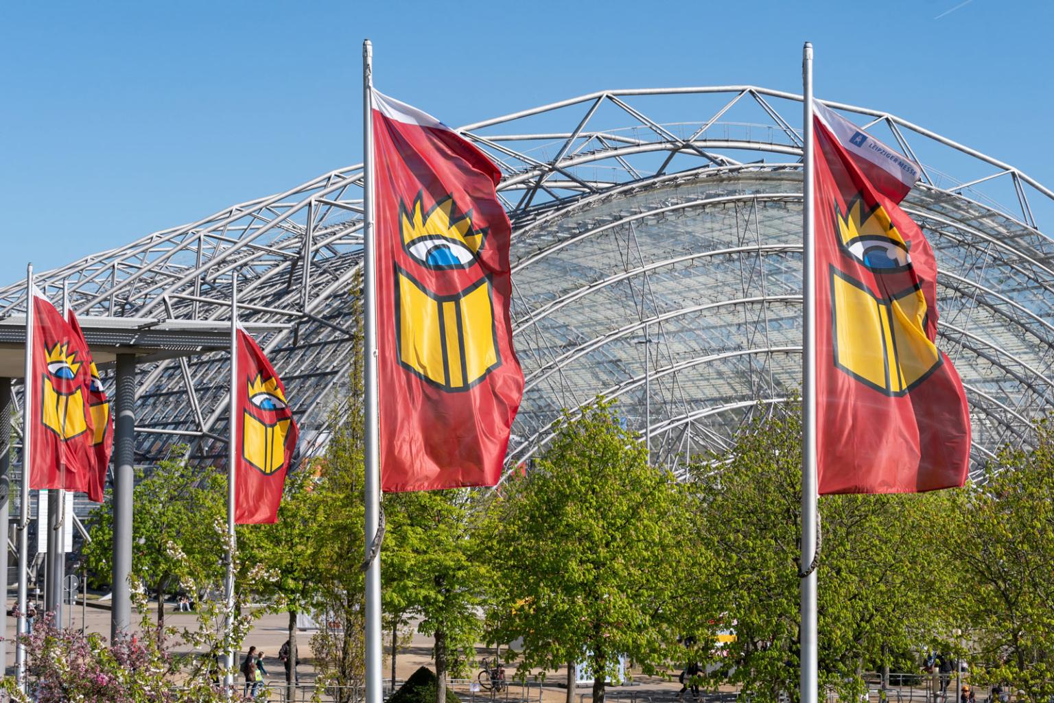 Vier Fahnen mit dem Motiv der Leipziger Buchmesse wehen im Wind vor einem großen Glasgebäude mit gewölbtem Dach. Die Fahnen zeigen ein Auge und ein aufgeschlagenes Buch.