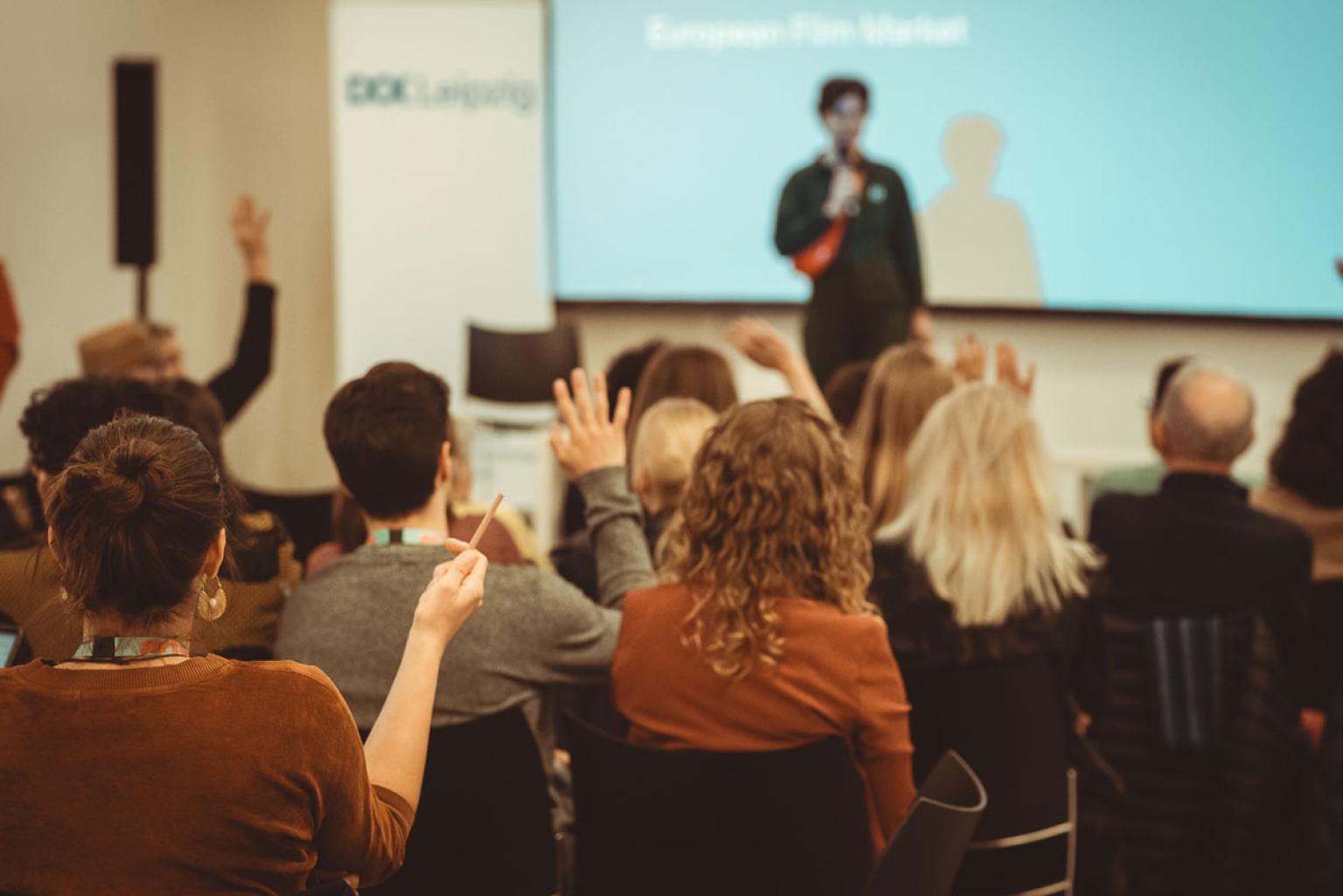 Several people sit in rows in a conference room, a speaker is on stage. Four persons in the audience raise their hands.