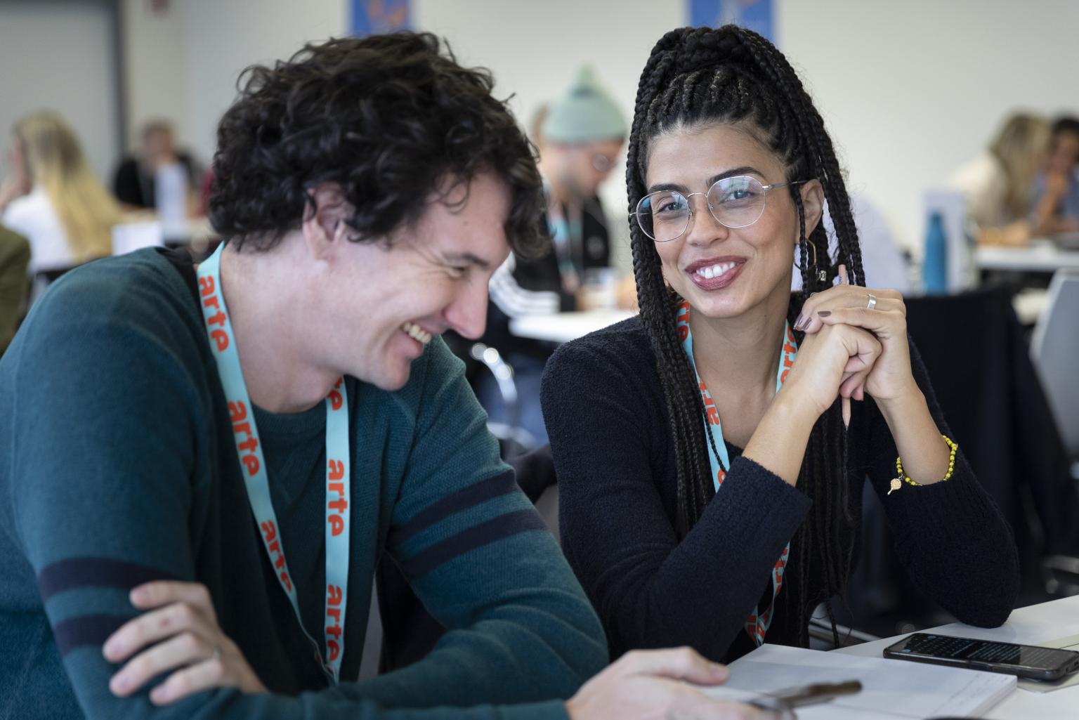 A woman of colour and a white man, both laughing, are sitting at a table at the Co-Pro Market