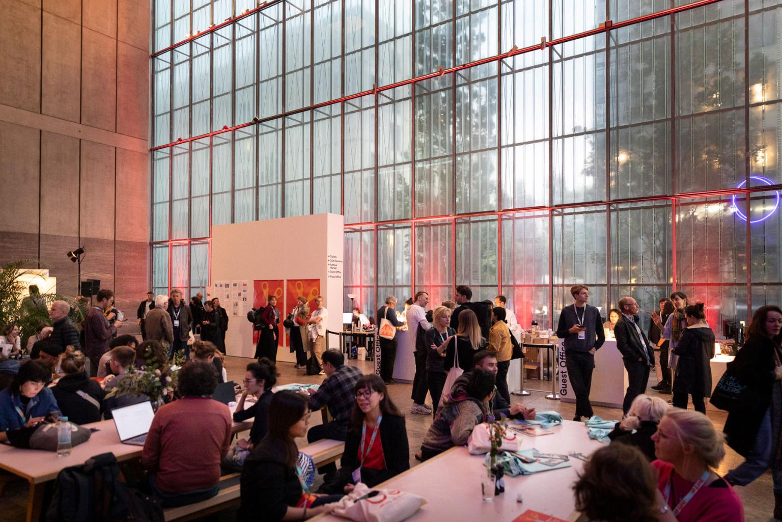 The festival centre, a large, bright room with a window front. Many people are busy sitting at tables or standing together, engaged in conversation.