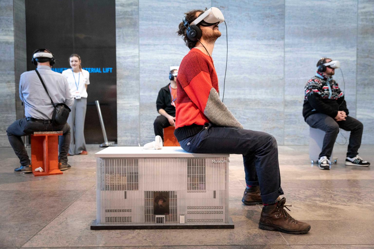 A man inside the museum of fine arts is wearing vr glasses while sitting on a miniature version of the museum of fine arts