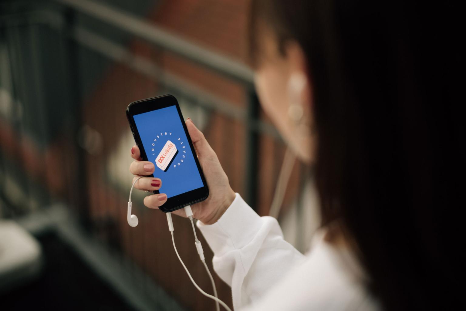 Eine Person hält ihr Handy und Kopfhörer hoch, auf dem Bildschirm leuchtet das Dok Industry Podcast Logo