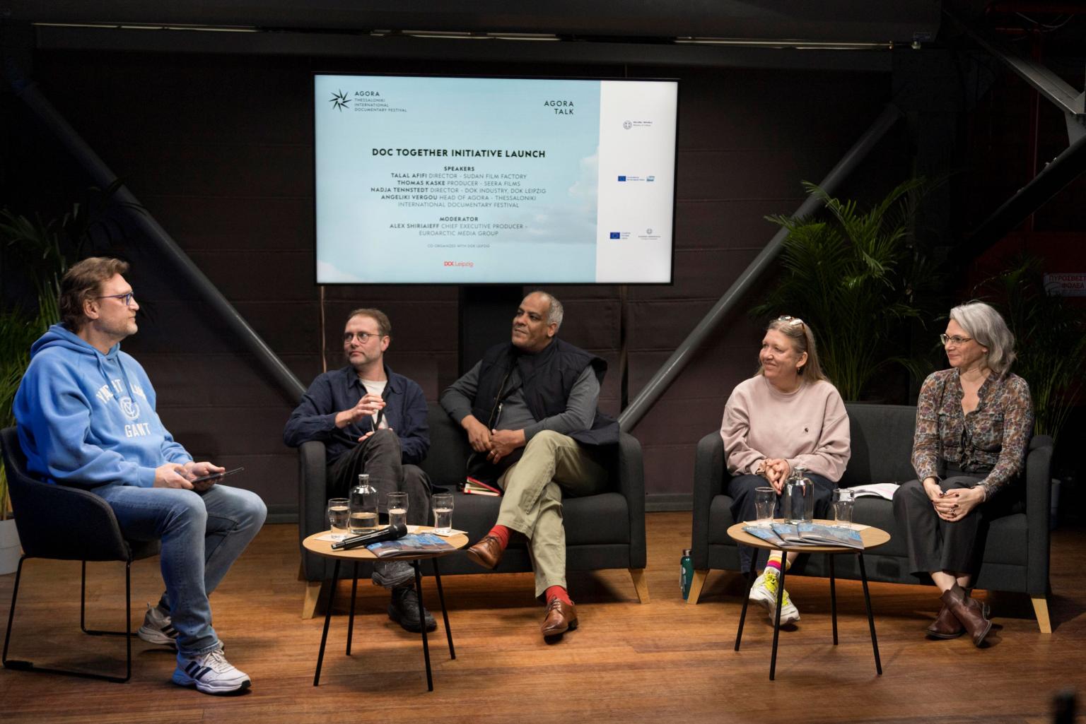 Three men and two women are sitting on a stage with a screen in the background displaying the topic "Doc Together Initiative Launch."