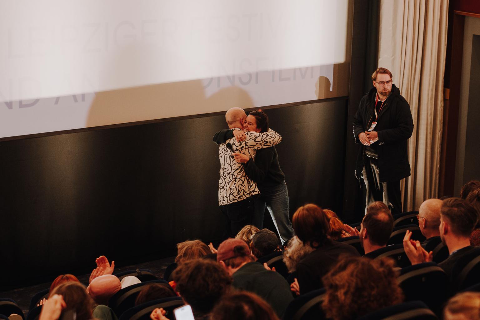 Vor einem Kinopublikum fallen sich der Regisseur Sandi DuBowski und Moderatorin Julia Weigl in die Arme. Die Leinwand hinter ihnen ist beleuchtet