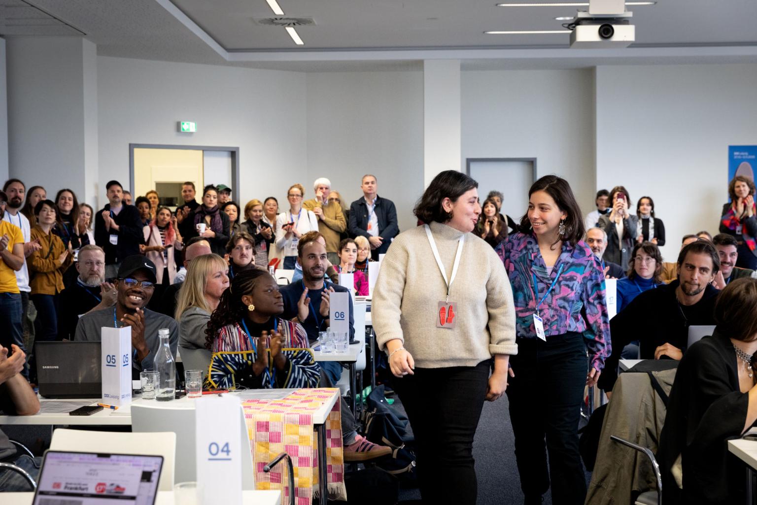 Two women are walking towards the camera in a crowded room, people are applauding