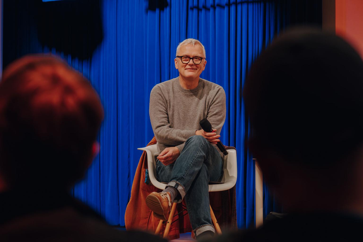 Man with gray hair and glasses sits smiling with a microphone in front of a blue curtain, audience in the foreground.