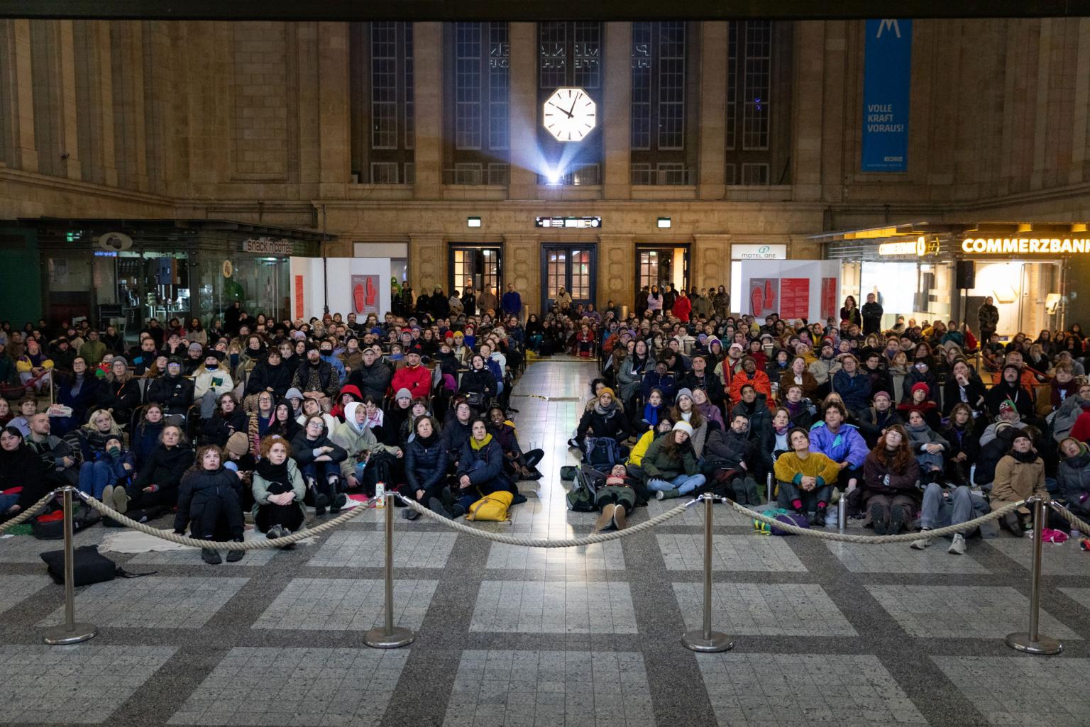 The eastern hall of the main station Leipzig is full of people, many of them sitting on chairs but also comfortably on the floor watching the big screen behind the camera