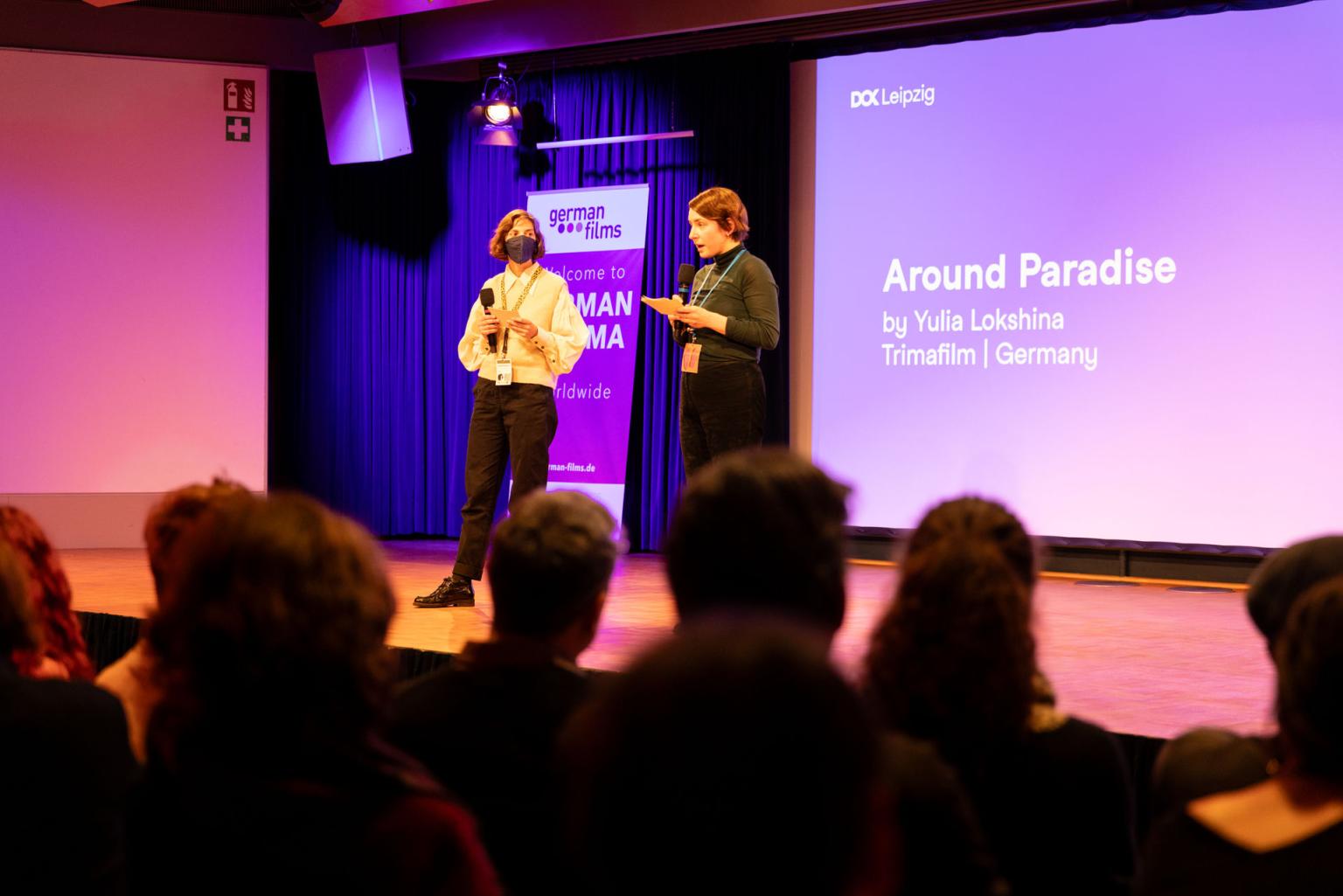 Two people are pitching on stage, several heads from the audience can be seen. Behind them the text "Around Paradise by Yulia Lokshina"