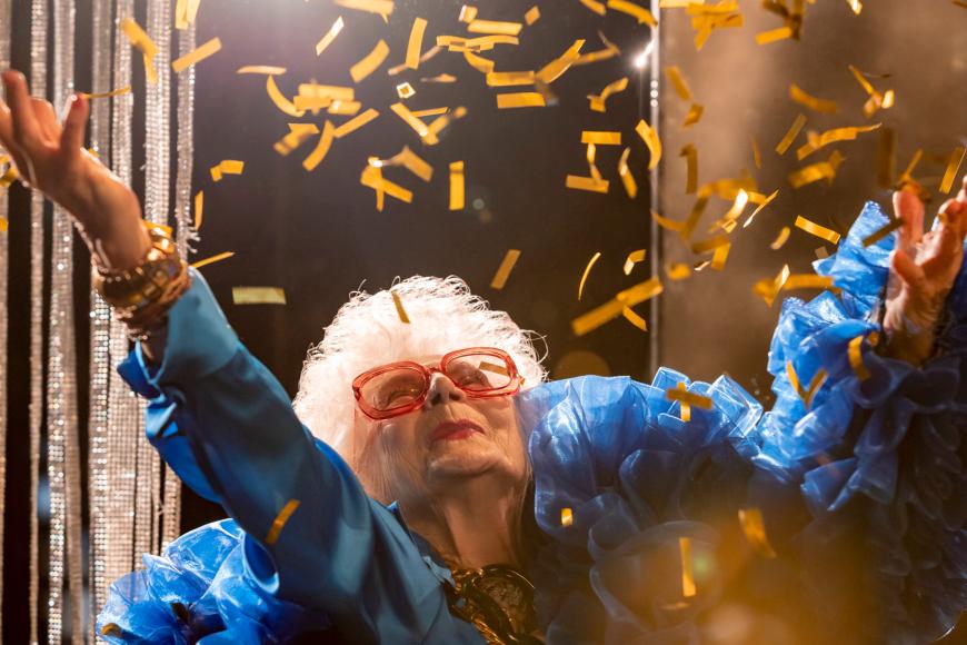 Eine alte Frau mit weißen Haaren und rotem Lippenstift in einem bauschigen blauen Kleid wirft goldenes Konfetti über sich