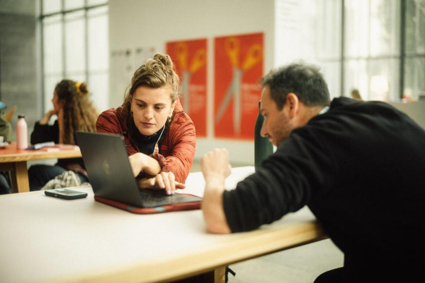 Two persons look at a laptop display while talking to each other.