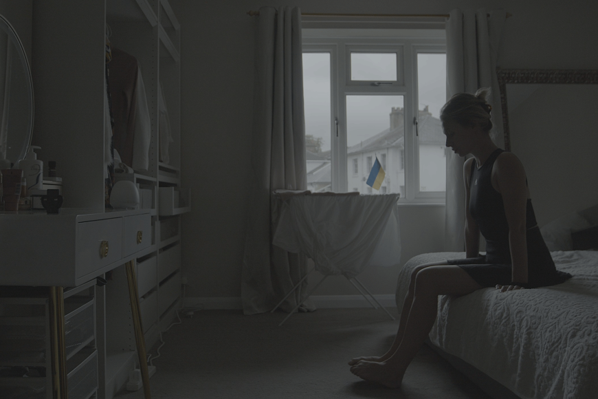 A woman sits on a bed in a room with white interior, in the window a Ukrainian flag.