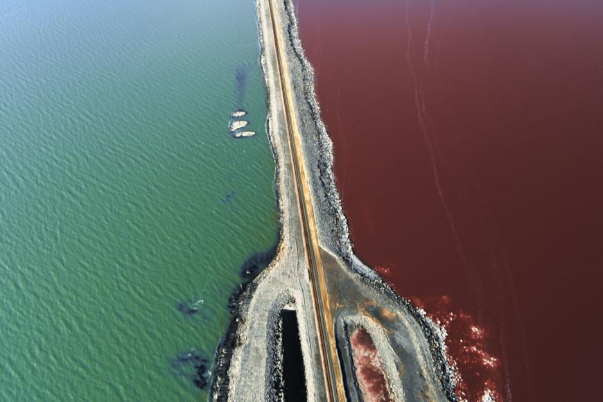 Eine leere Straße führt zwischen zwei Gewässern hindurch. Das Wasser links ist bläulich, das Wasser rechts der Straße rot-braun.