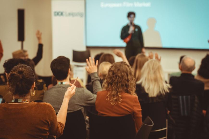 Several people sit in rows in a conference room, a speaker is on stage. Four persons in the audience raise their hands.