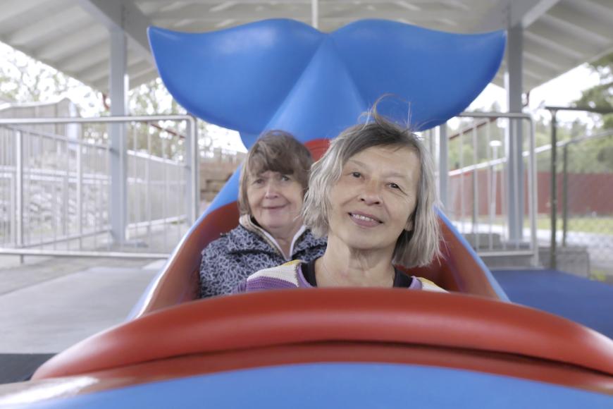 Zwei ältere Frauen sitzen in einem offenen Wagen von einer Spielzeuggefährt in Form eines Wals.