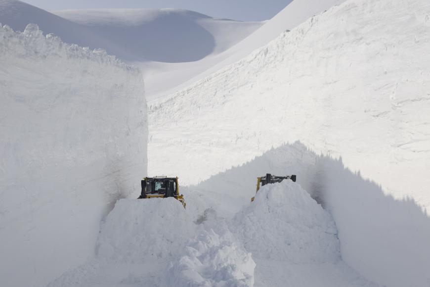 Zwei Schneeraupen räumen eine tiefe, schmale Schneise durch meterhohen Schnee in einer verschneiten Berglandschaft.