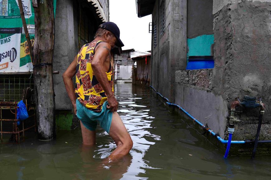 Ein Mann watet in kniehohem Wasser, das eine Straße zwischen engstehenden Häusern geflutet hat.